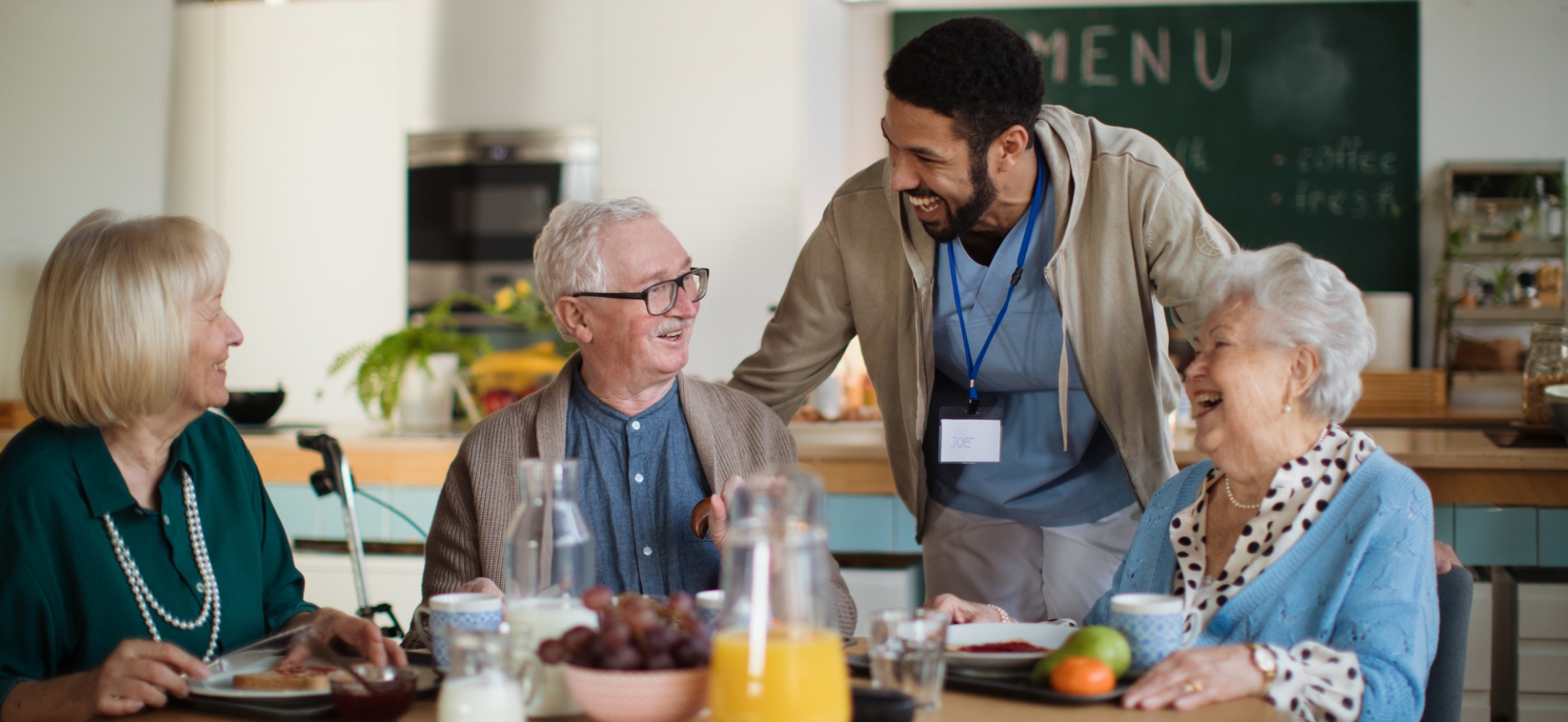 elderly people sitting around a table having breakfast. Male nurse laughing and in conversation with the elderly people.